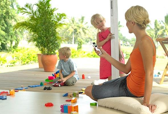 Eine Frau sitzt im Wohnzimmer auf einem Sitzkissen vor der offenen Terrassentür und hält eine Fernbedienung in der Hand. Im Hintergrund sieht man spielende Kinder.