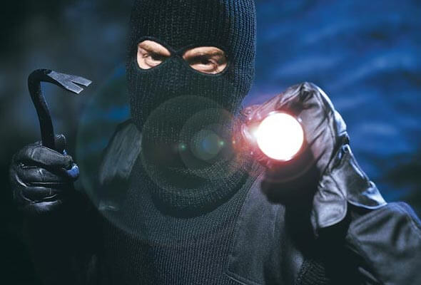 Ein Einbrecher mit schwarzer Maske hält in der einen Hand ein Brecheisen und in der anderen Hand eine Taschenlampe